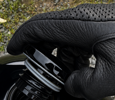A close-up of a black leather motorcycle glove being fastened onto a black motorcycle fuel tank, with gravel and grass visible in the background.