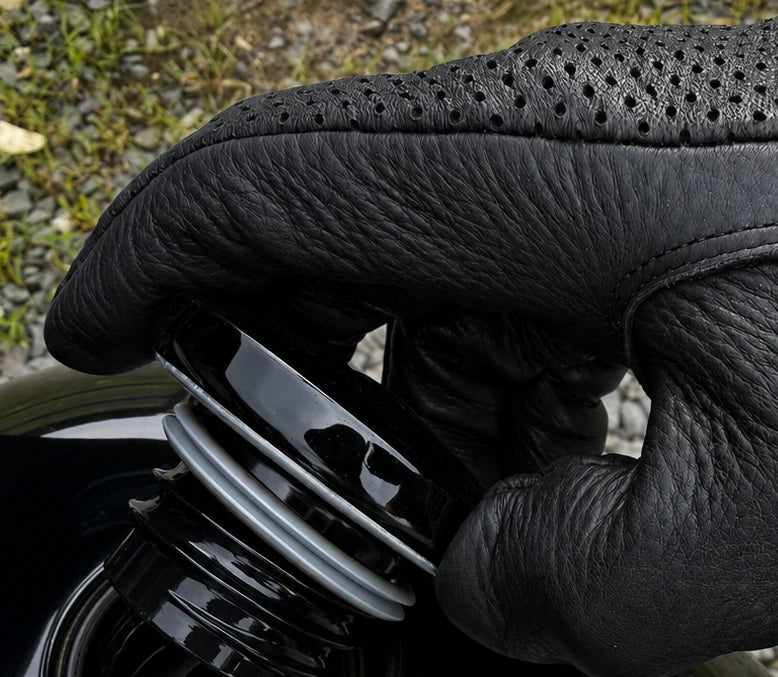 A close-up of a black leather motorcycle glove being fastened onto a black motorcycle fuel tank, with gravel and grass visible in the background.