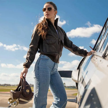 Woman wearing Cockpit USA Women's Amelia leather jacket standing on aircraft wing holding flight helmet