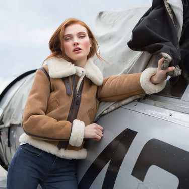 Woman modeling the tan suede and sheepskin Cockpit USA Women’s Hooded B-3 Bomber Jacket next to an aircraft